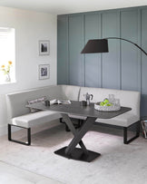 Modern dining nook featuring a white leather upholstered L-shaped bench and a rectangular dining table with a sleek black top and an X-shaped metallic base. A floor lamp with a curved stand and large black shade extends over the table.