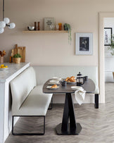 A contemporary dining area featuring an oval table with a matte black pedestal base, complemented by a cream upholstered corner bench with a sleek black metal frame.