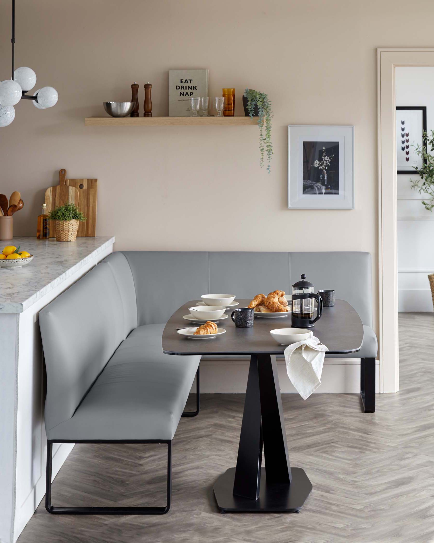 Modern minimalist dining area with a grey upholstered corner bench featuring a sleek black metal base, paired with a contemporary round table with a dark grey tabletop and a bold geometric black pedestal base.