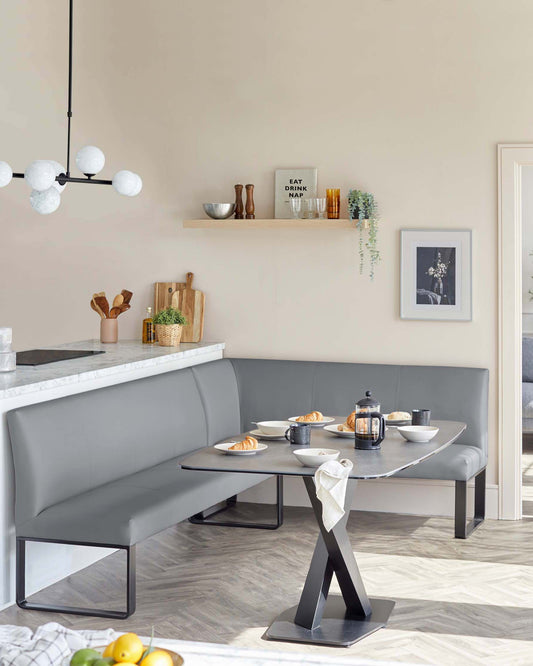 Modern corner dining nook with a grey upholstered bench seating and a rectangular table featuring a sleek dark metal base and a grey stone-like top. A wooden floating shelf is mounted above the bench.