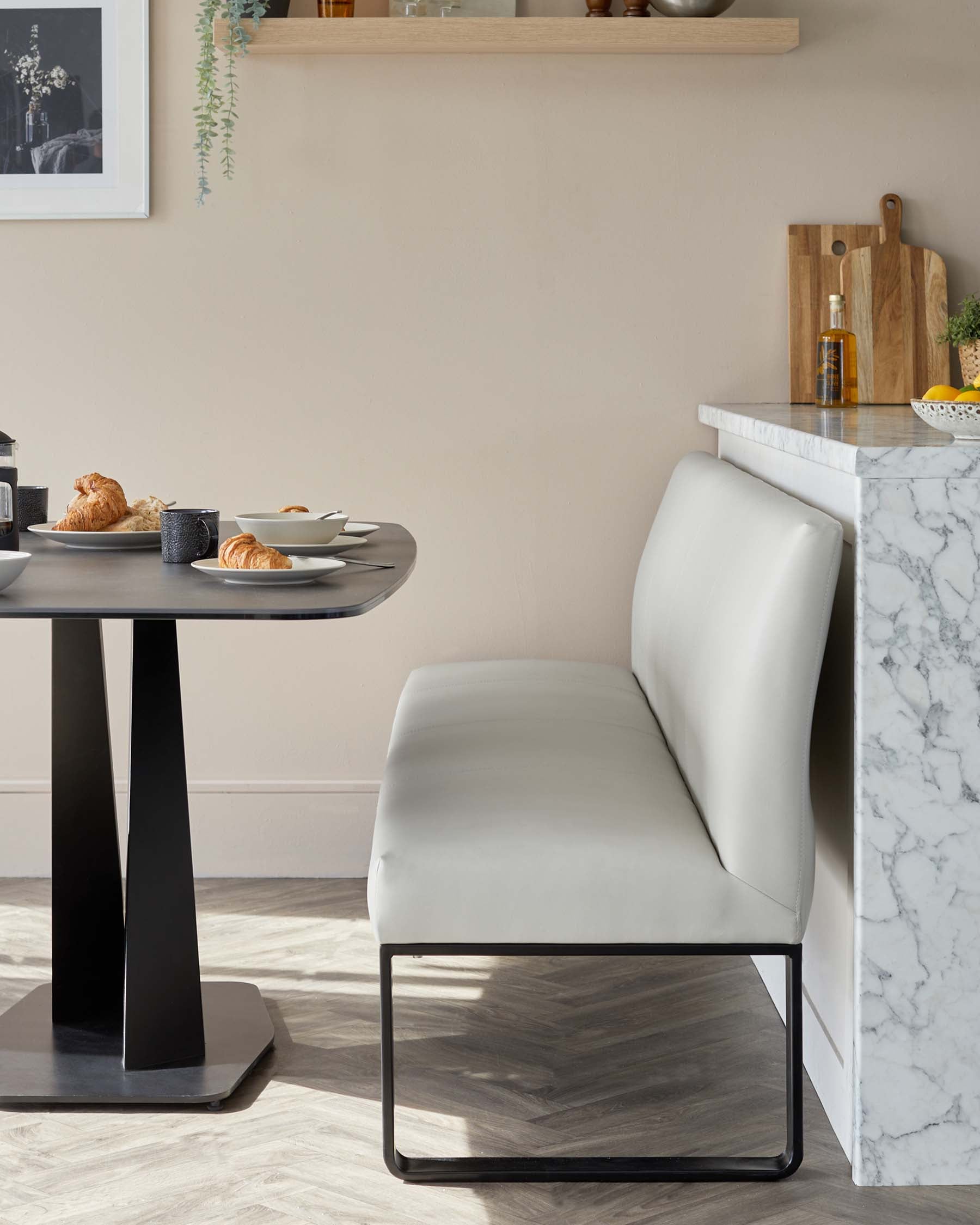 Modern dining setting with a round, dark-finished table with a unique central pedestal base paired with a sleek, light-coloured upholstered bench with a minimalist black metal frame. A marble-top kitchen counter adds a touch of luxury in the background.