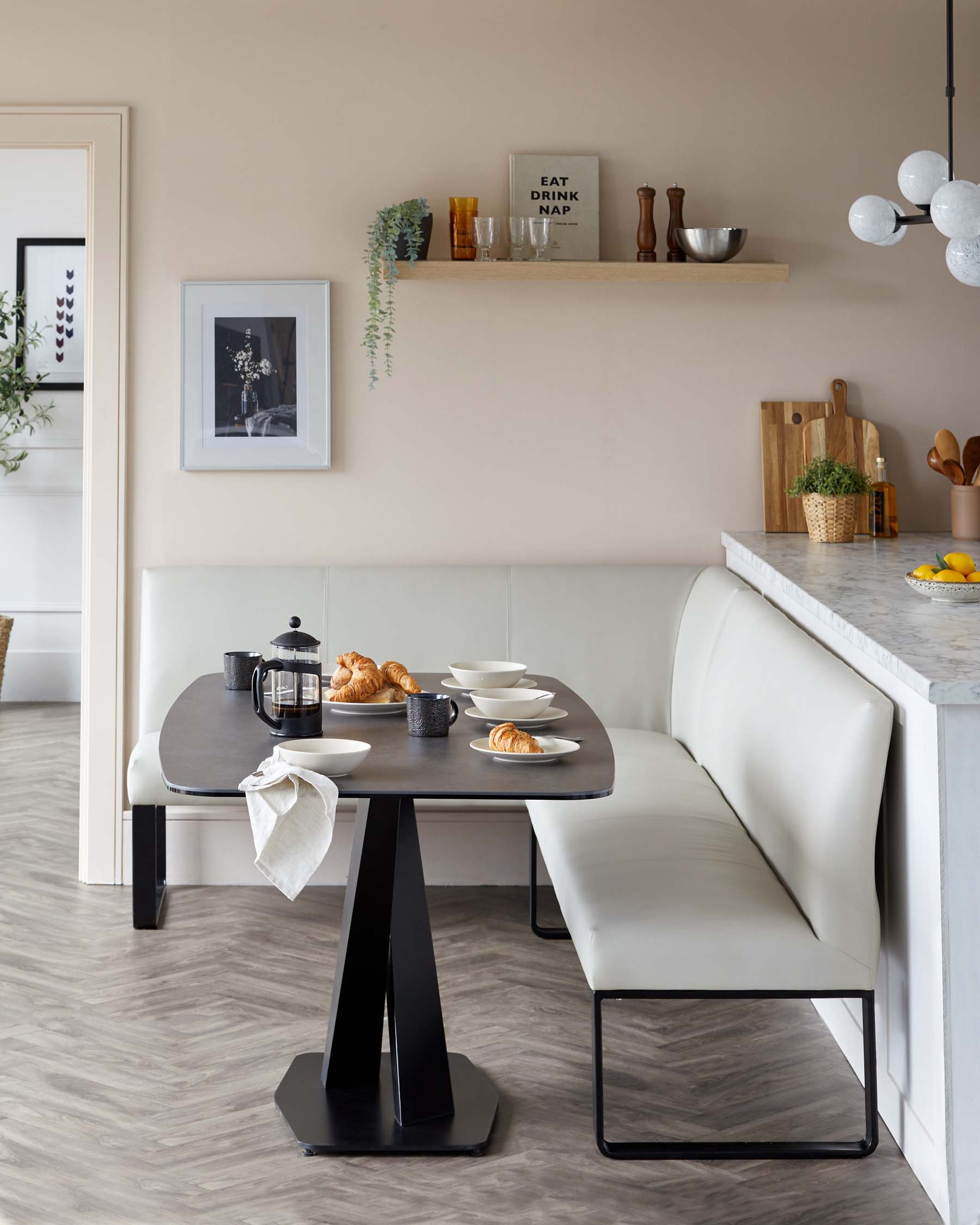 Modern dining area featuring a sleek, black oval table with a unique geometric base design. Accompanied by a contemporary, cream-colored corner bench seat with a minimalist frame and a matching single chair with black metal legs.