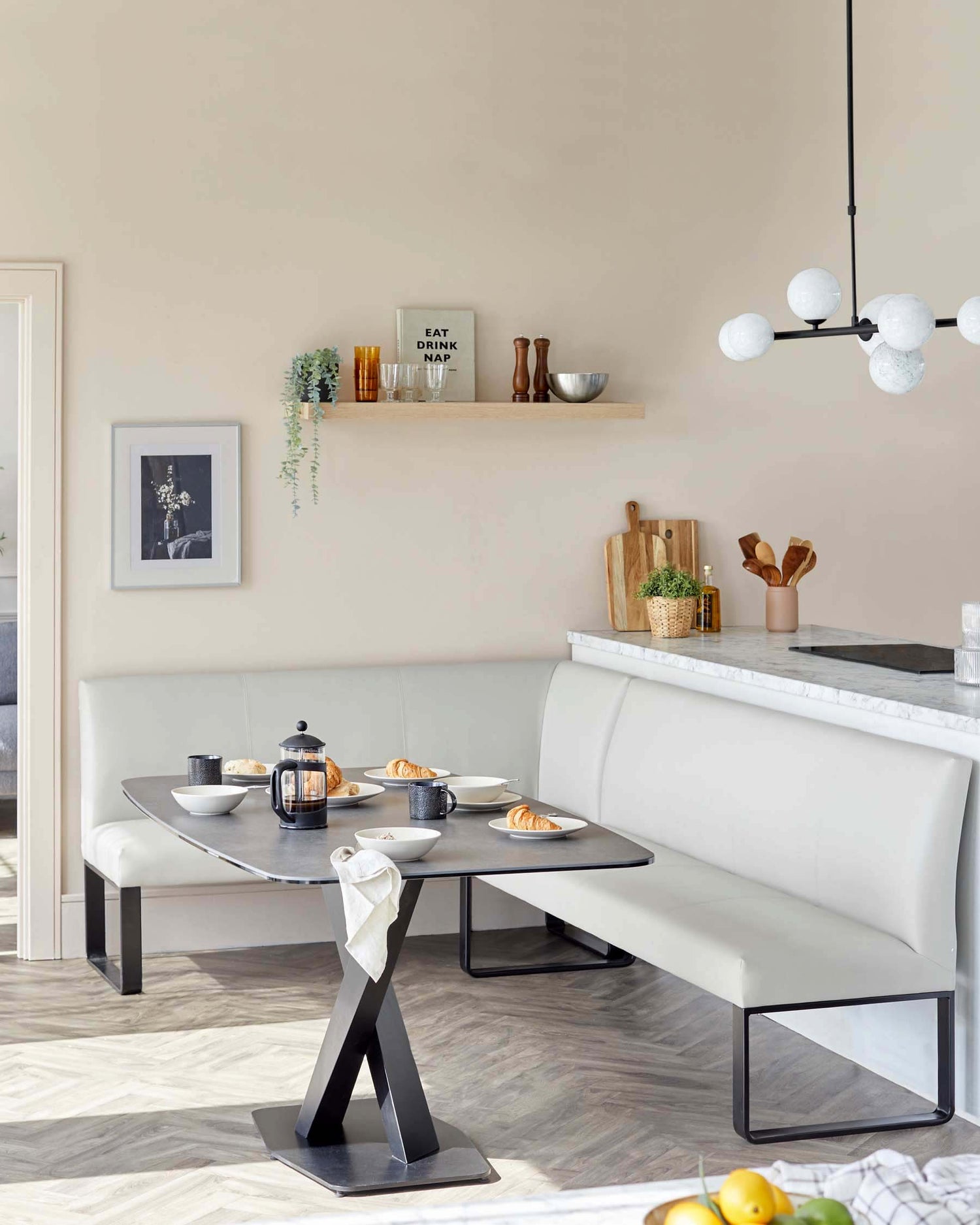 Modern dining setting featuring a sleek, rectangular table with a dark top and a unique, central black metal pedestal base in a sculptural 'A' shape. The dining area is completed with an L-shaped, cream-colored bench with a padded backrest and seat, designed to seamlessly blend comfort with contemporary aesthetic. The furniture is complemented by a minimalist kitchen counter with a marble top in the background.