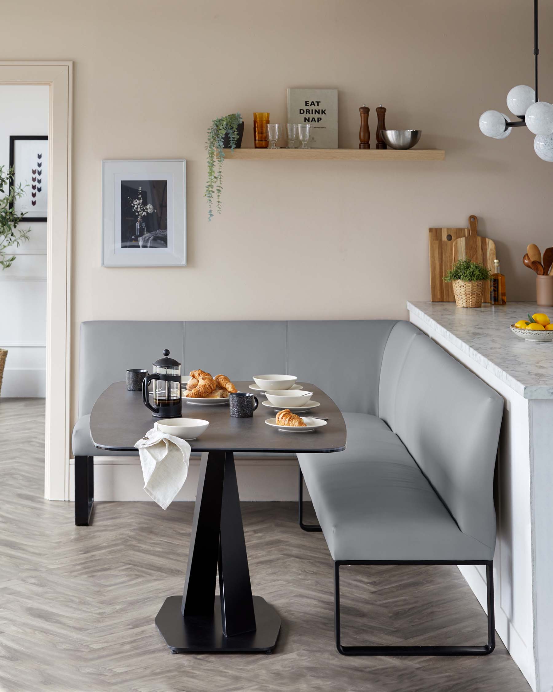 Modern dining area featuring a dark, rectangular table with a unique central pedestal base in black. Accompanying the table is a corner bench upholstered in a sleek, grey material with a minimalist metal frame, offering a streamlined and contemporary seating solution.