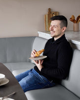 Modern minimalist dining nook with a built-in light grey leatherette bench featuring vertical stitching, complemented by a white marble tabletop with a smooth finish. There's a set of wooden cutting boards as wall decor adding a natural accent to the space.