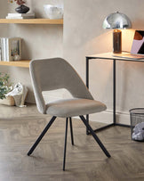 Modern upholstered chair with a smooth light grey fabric and a sleek curved backrest, featuring angled black metal legs. A minimalist black frame desk with a light wooden top, accompanied by a chrome dome table lamp and decorative objects, is partially visible in the background.