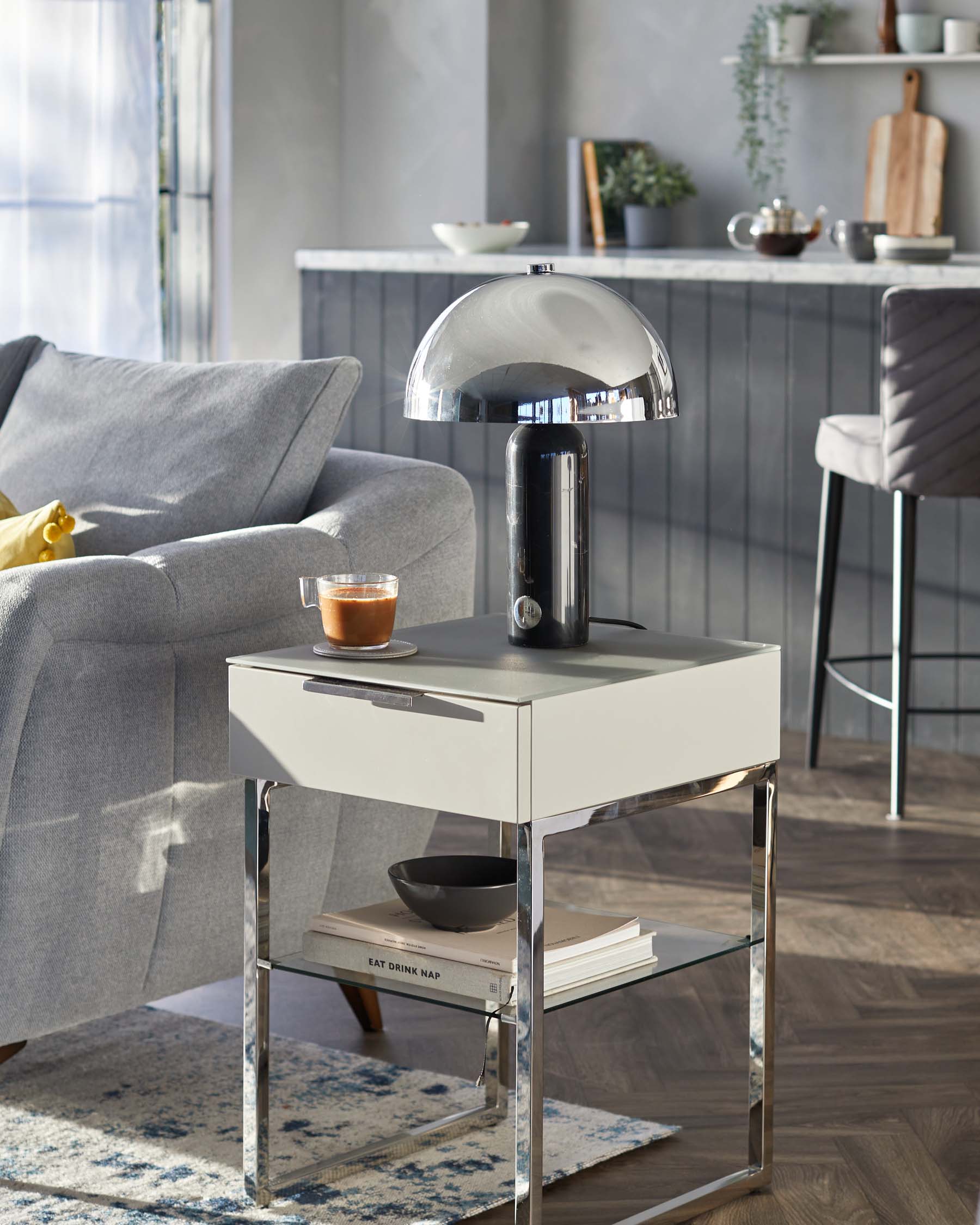 Modern side table with a sleek grey top and mirrored chrome legs, featuring an open lower shelf with stacked books, alongside a grey fabric sofa and a reflective metallic desk lamp.