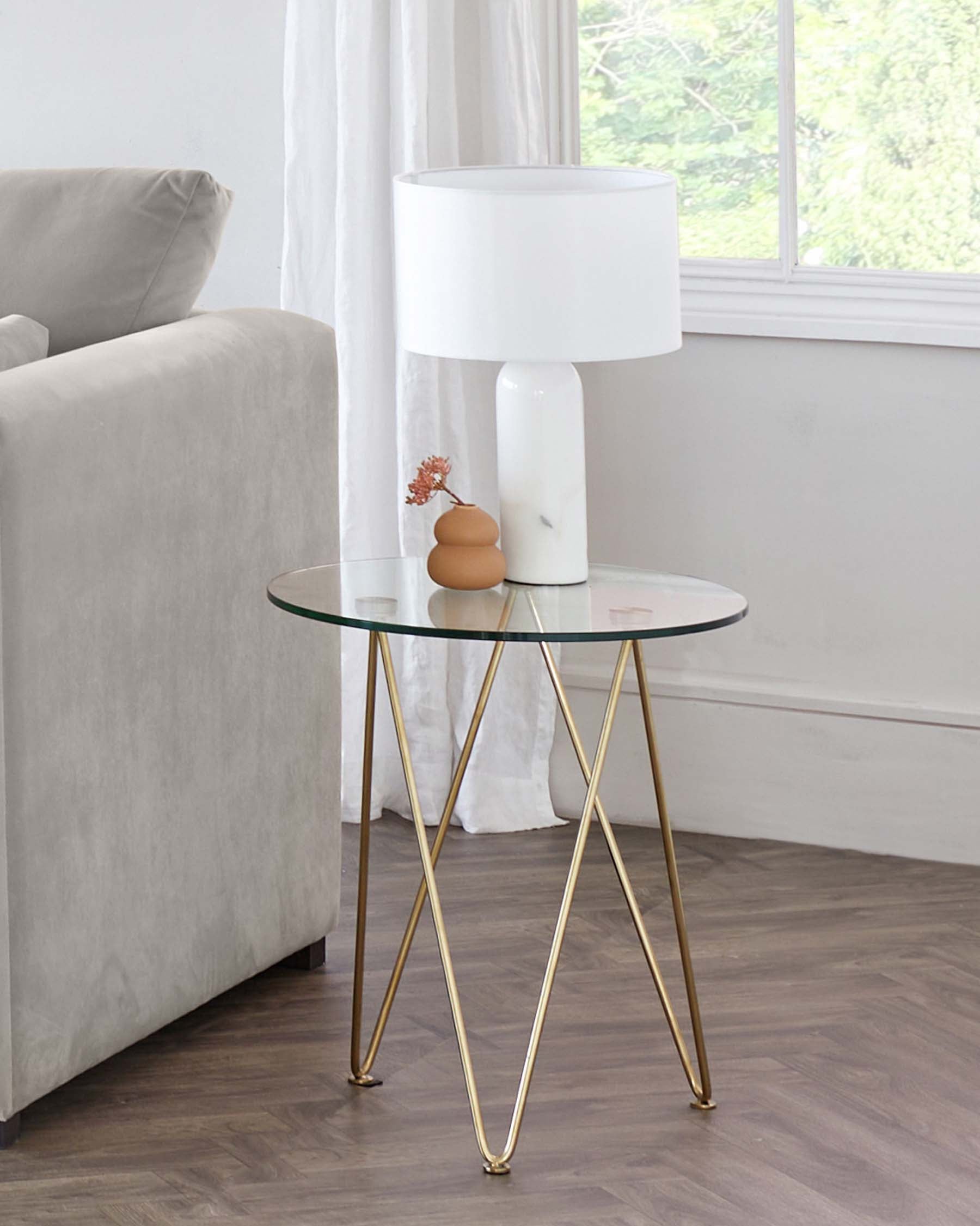 Round glass-top side table with slender gold-finished metal legs in an X-frame design, positioned next to a light grey sofa, with a large white lamp and small decorative items atop, in a room with a window, white curtains, and wooden flooring.