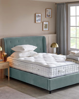 A soft, tufted teal bed frame with a plush white mattress, layered with pillows, and a bedside table featuring a flower vase.