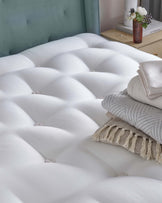 A plush white mattress with tufted detailing, layered with neatly folded blankets and decorative cushions against a soft green headboard.