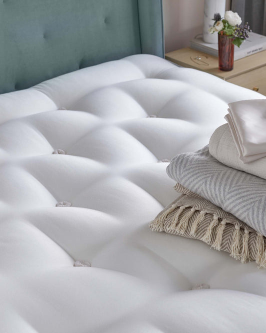 A plush white tufted mattress with neatly stacked light-colored blankets and a cozy throw, set against a soft blue headboard.