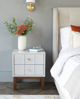 Modern nightstand with two drawers, wooden legs, a decorative vase with flowers, and a book with glasses on top.