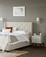 Cozy bedroom featuring a tufted beige bed, elegant nightstand, soft throws, and stylish wall sconces against a muted backdrop.