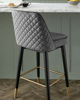 Elegant modern bar stool with a quilted, diamond-pattern backrest and a smooth seat in a grey fabric upholstery. The chair features four tapered legs in a black finish with sleek brass-coloured footrests and matching brass tips on the legs, offering a luxe, contemporary aesthetic.