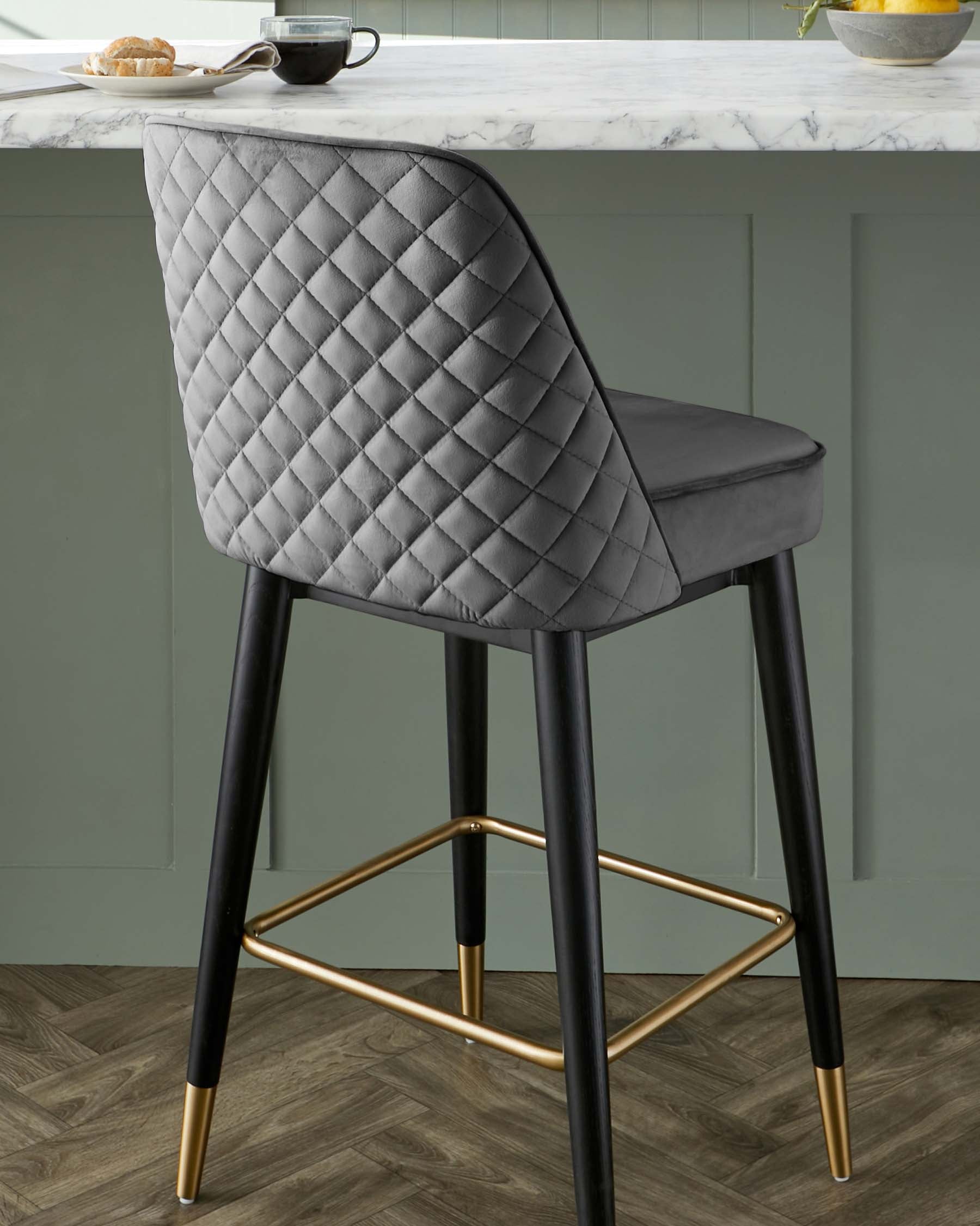 Elegant modern bar stool with a quilted, diamond-pattern backrest and a smooth seat in a grey fabric upholstery. The chair features four tapered legs in a black finish with sleek brass-coloured footrests and matching brass tips on the legs, offering a luxe, contemporary aesthetic.