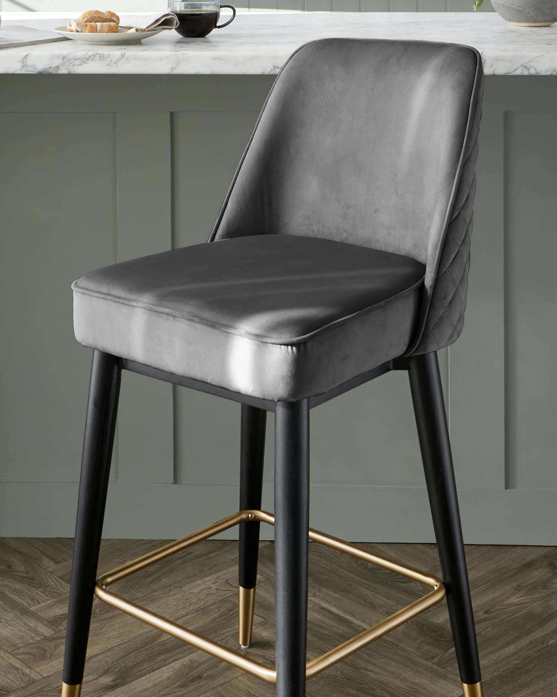 Elegant modern bar stool with a luxurious grey velvet upholstered seat and backrest featuring diamond tufting details. The chair has sleek black tapered legs with gold accents at the feet and footrest, offering a stylish contrast and a sophisticated touch.