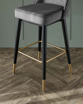 Elegant modern bar stool with a grey upholstered seat and backrest featuring quilted detailing. The stool has black tapered legs complemented by gold-tone footrests and feet accents. The piece is set against a herringbone-pattern wooden floor and a green wall, suggesting a contemporary style with a touch of luxury.