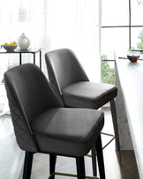 Two elegant modern dining chairs with dark grey upholstery and a quilted backrest design. The chairs have a sleek silhouette with slightly flared legs and footrests in a matte black finish with metallic accents. The setting is complemented by a glimpse of a white marble dining table and a side table with decorative items, all bathed in natural light from a window with sheer curtains.