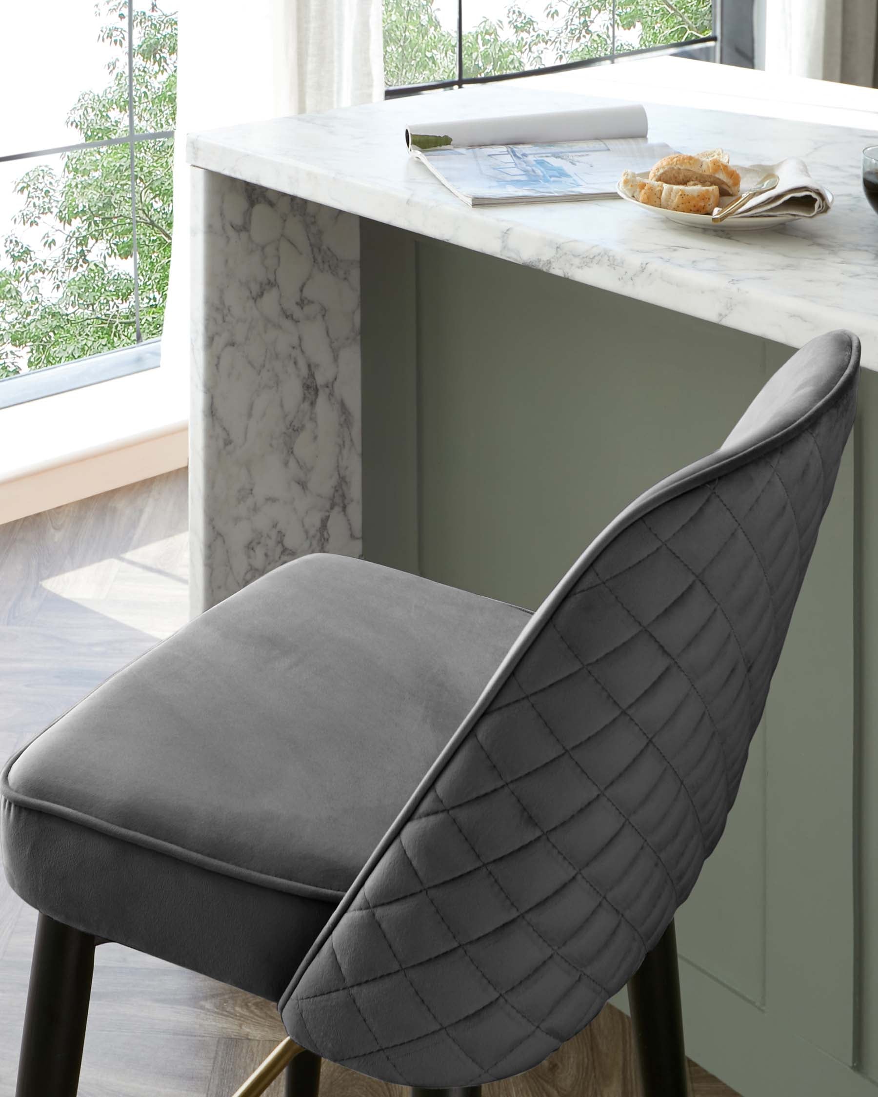 Modern grey upholstered dining chair with quilted backrest detail and slim metallic legs, paired with a white marble kitchen island.