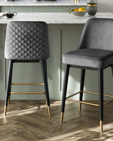 Two modern bar stools with dark grey upholstery and diamond tufting detail on the backrest. The stools have slender black metal legs with golden accents at the feet and bracing bar.