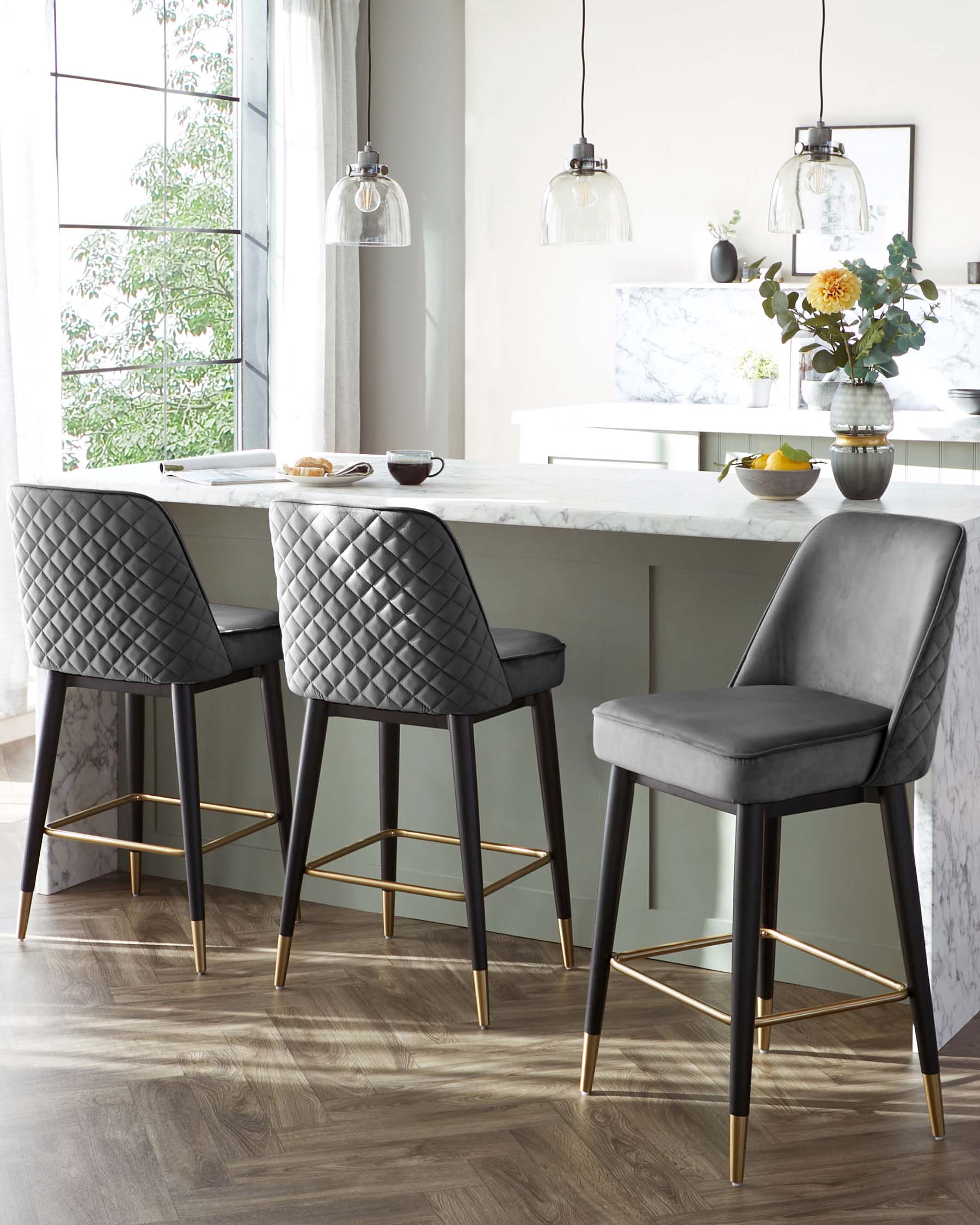 Elegant modern bar stools with dark grey upholstered seats and backs featuring a diamond tufting design. The stools have sleek wooden legs in a dark finish, accented with gold footrests, complementing a white marble countertop in a contemporary kitchen setting.