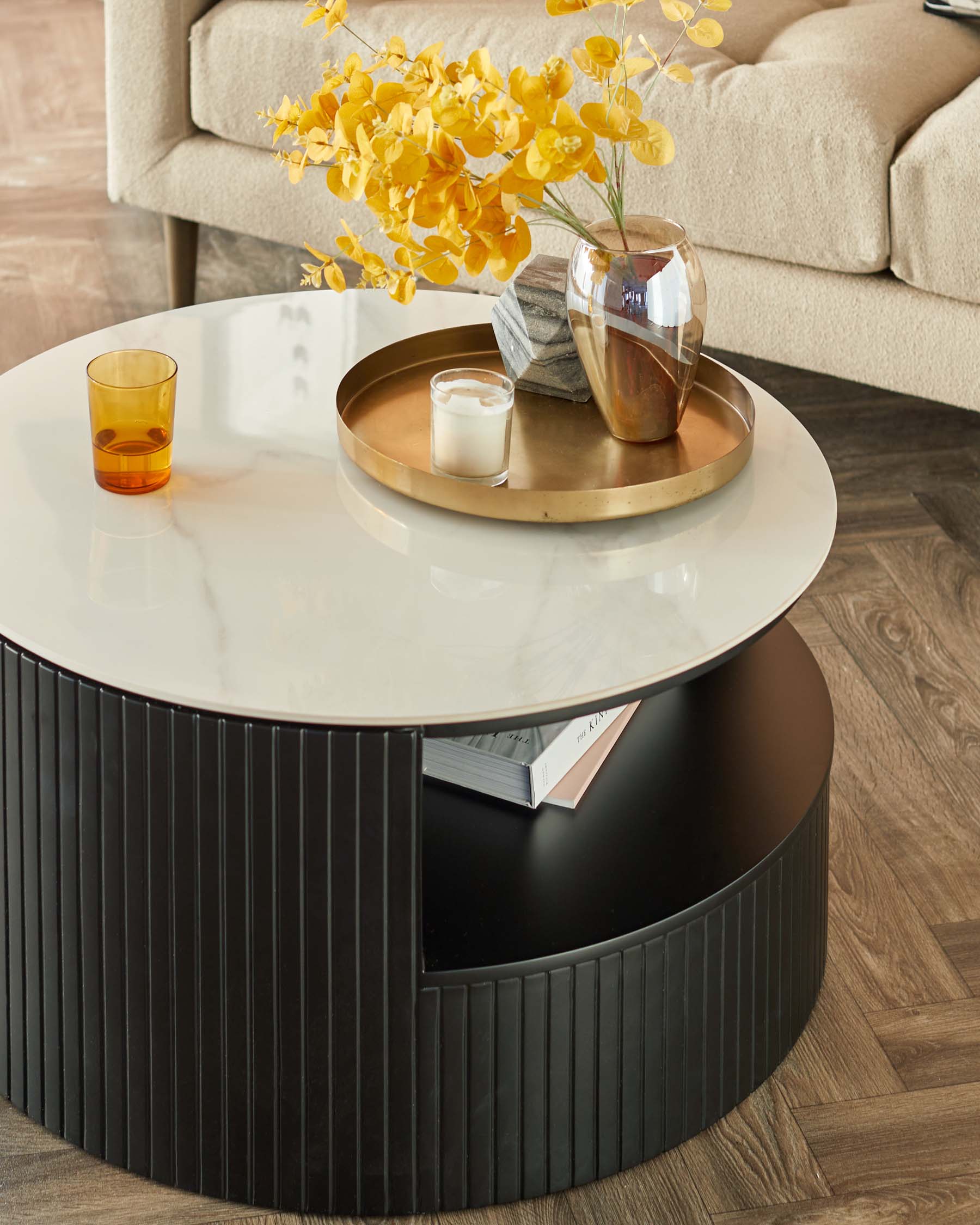 Round modern coffee table with a glossy white tabletop and a ribbed black base, accessorized with a brass tray, a vase with yellow flowers, a candle, and a glass. A beige sofa with textured upholstery is partially visible in the background.