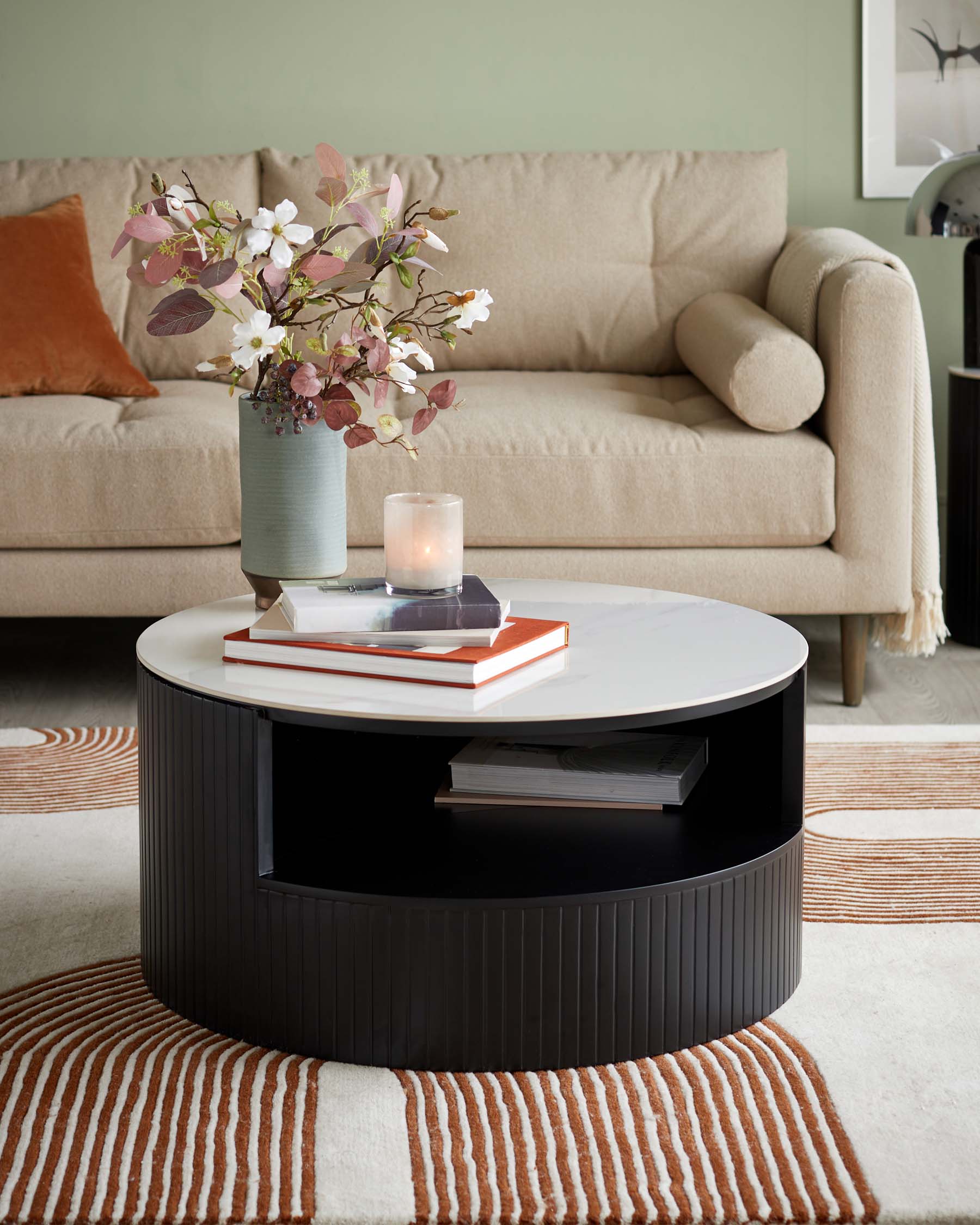 A contemporary beige fabric sofa with plush cushions and a cylindrical bolster pillow at one end, paired with a round, two-tiered coffee table featuring a white top and a dark lower section with vertical fluting and an open shelf for books. The setting is complemented by a striped area rug in warm earth tones.