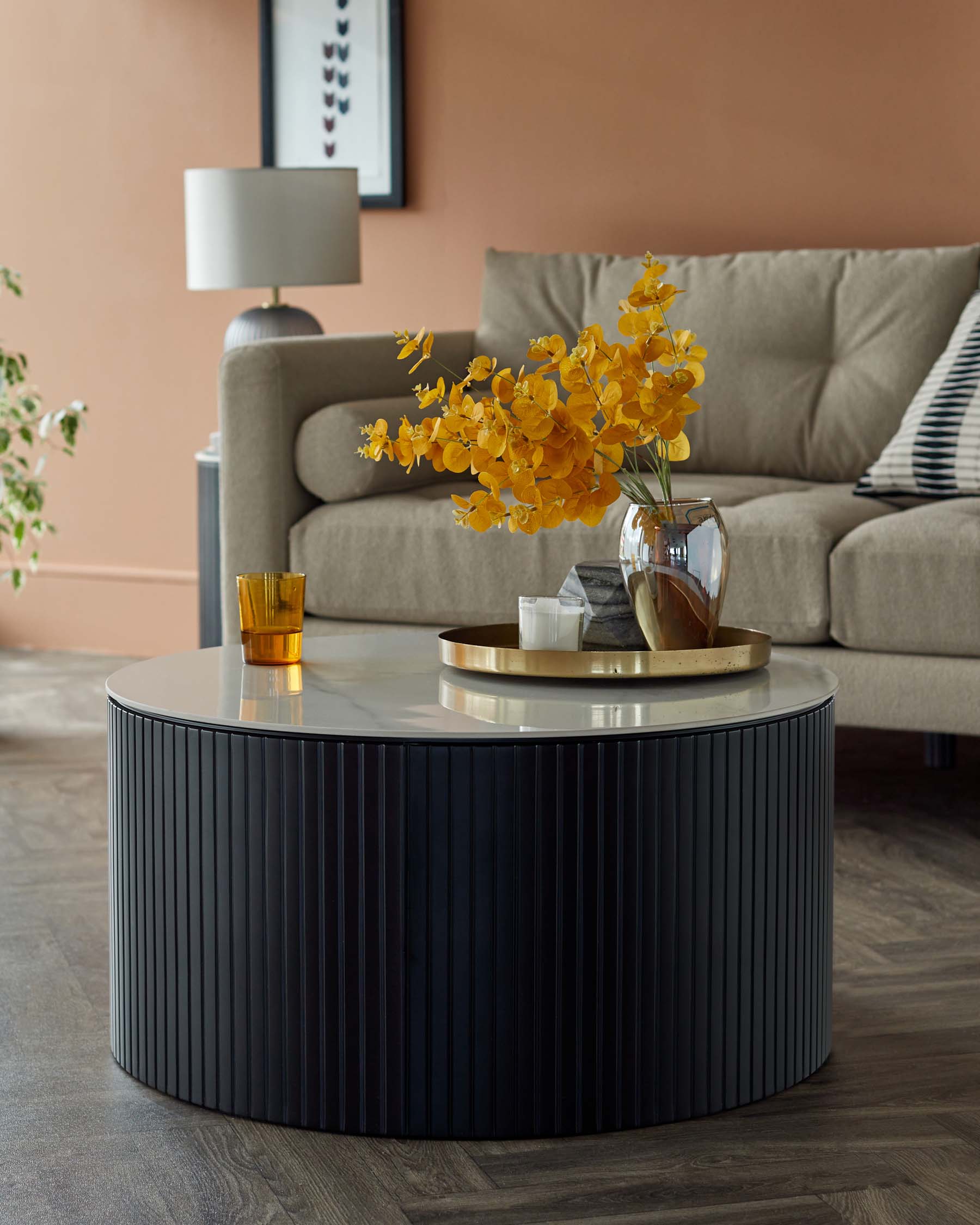 A modern round coffee table with a sleek, black ribbed base and a reflective metallic top, paired with a minimalist grey fabric sofa. There's also a white table lamp with a circular base and a cylindrical shade in the back.