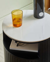 A modern round side table with a white marble top and a fluted black base, featuring a book on the lower shelf and decorations on top.