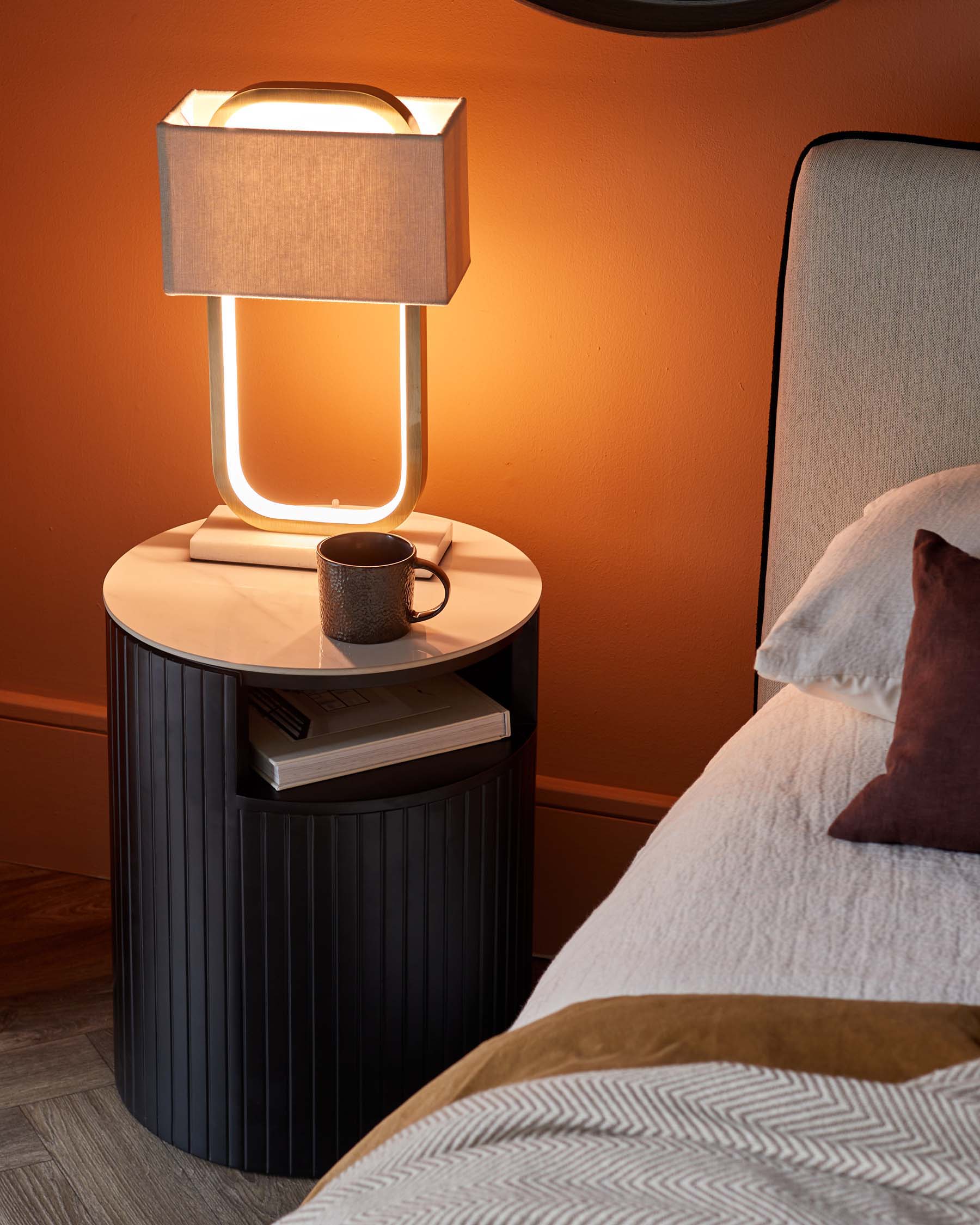 Modern cylindrical bedside table with vertical fluting and a white marble top, paired with an avant-garde table lamp featuring a rectangular fabric shade and a unique illuminated frame.