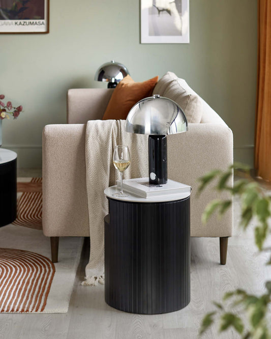 A contemporary living room setup with a beige, upholstered armchair featuring a textured throw and a brown decorative cushion. Adjacent to it is a round black side table with vertical fluting, which is styled with a sleek, silver dome table lamp, a book, and a glass of white wine. The furniture rests on a cream rug with orange zigzag patterns.