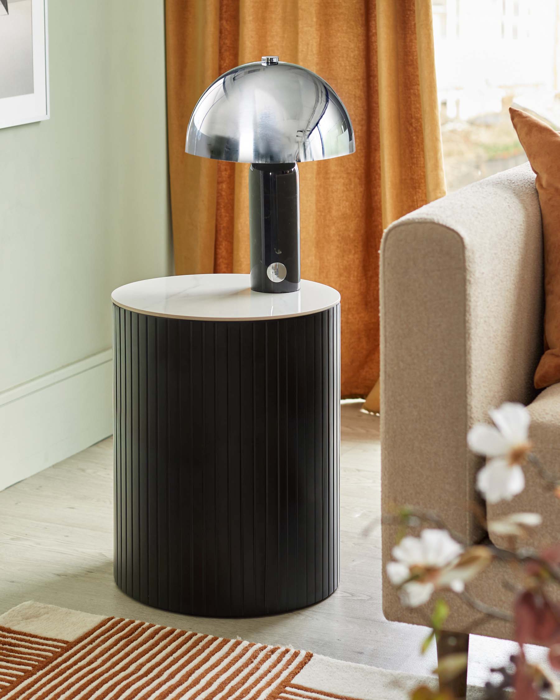 A modern cylindrical side table with a black vertical ribbed design and a white marble top, next to a beige sofa with a chrome mushroom-style lamp placed on it. The scene is complemented by a striped rug in earth tones on a hardwood floor.