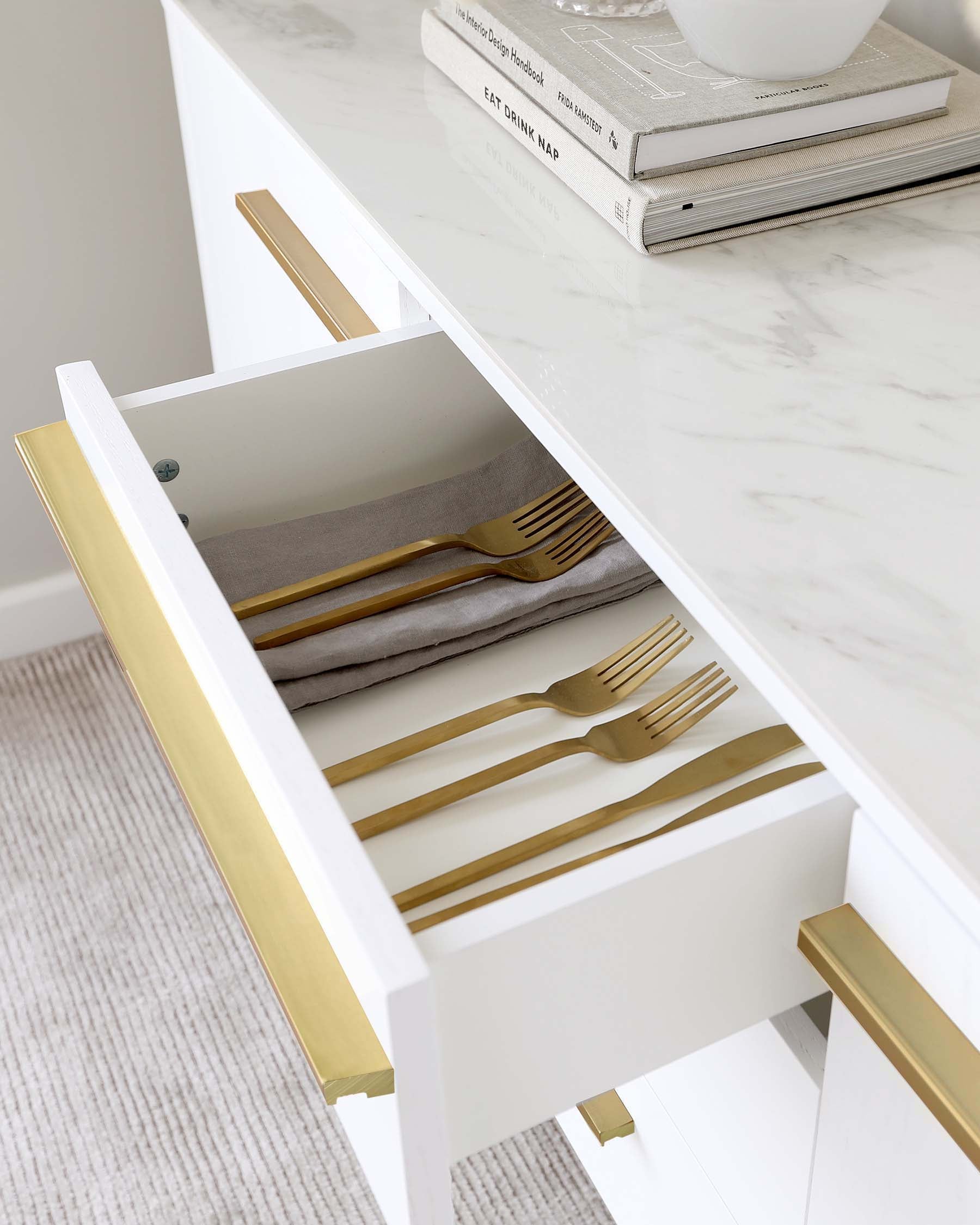 Elegant white console table with a marble top, showcasing gold handles on its open drawer and gold-tipped legs. The drawer interior features a neutral lining with neatly arranged cutlery inside. A stack of books sits on the table's surface, enhancing the minimalist aesthetic.