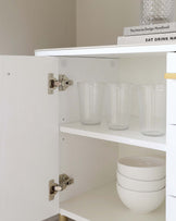 White open-faced wooden cabinet with two adjustable shelves displaying glassware and stacked bowls, featuring visible door hinges and a partial view of a book on top.