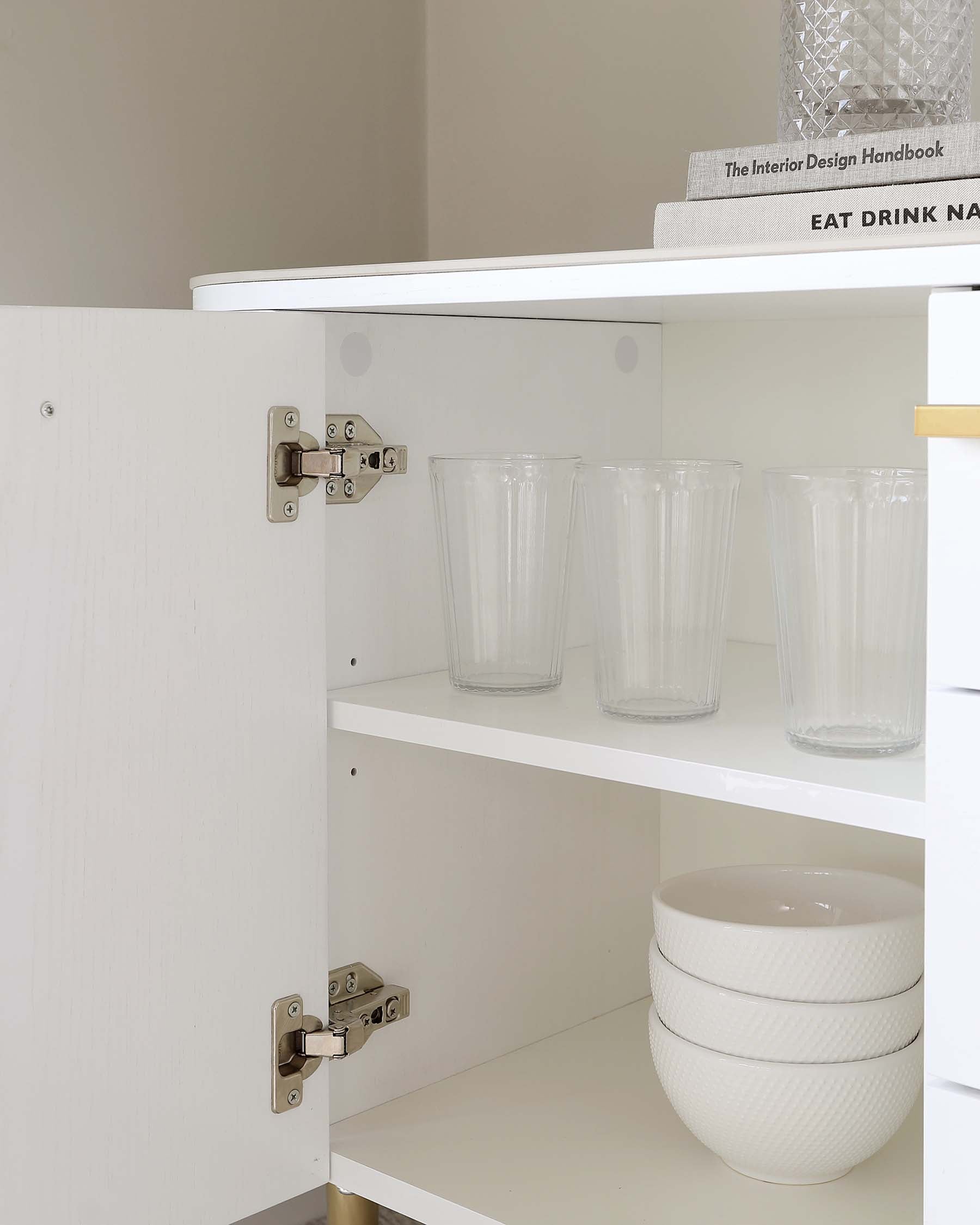 White open-faced wooden cabinet with two adjustable shelves displaying glassware and stacked bowls, featuring visible door hinges and a partial view of a book on top.