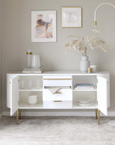 Modern white sideboard with gold accented legs and handles, featuring two hinged cabinet doors and a centre drawer, styled with decorative items such as a lamp, vase with dried pampas grass, books, and framed artwork above.