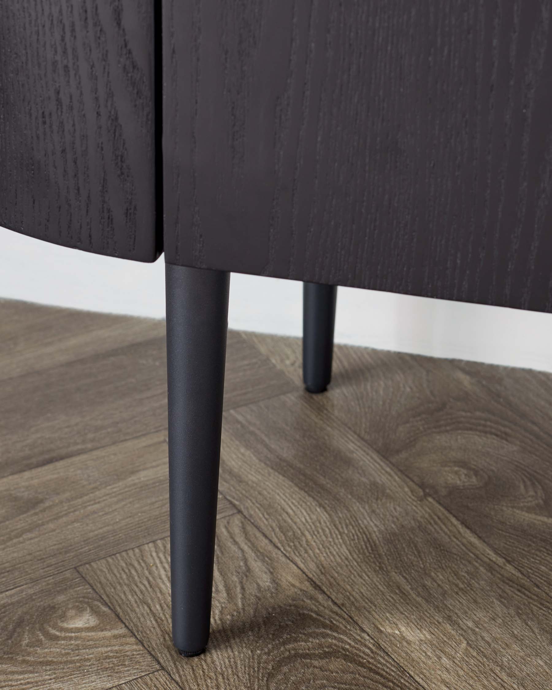 Close-up of a contemporary furniture piece with dark wood texture and elegant, sleek black legs, displayed on wooden flooring.