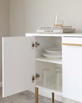 Modern minimalist white storage cabinet with open shelf displaying books and a glass, alongside an open door revealing an interior shelf with white plates and clear glasses. The cabinet features golden legs for a touch of elegance.