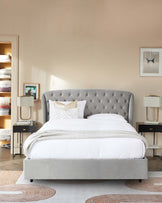 Elegant bedroom furniture set including a grey upholstered king-size bed with a tufted headboard, white bedding with decorative pillows, flanked by two black bedside tables with matching table lamps, set against a neutral wall with minimalist art and adjacent open wardrobe filled with neatly arranged bed linens. A stylish area rug with earthy tones adds warmth to the room.