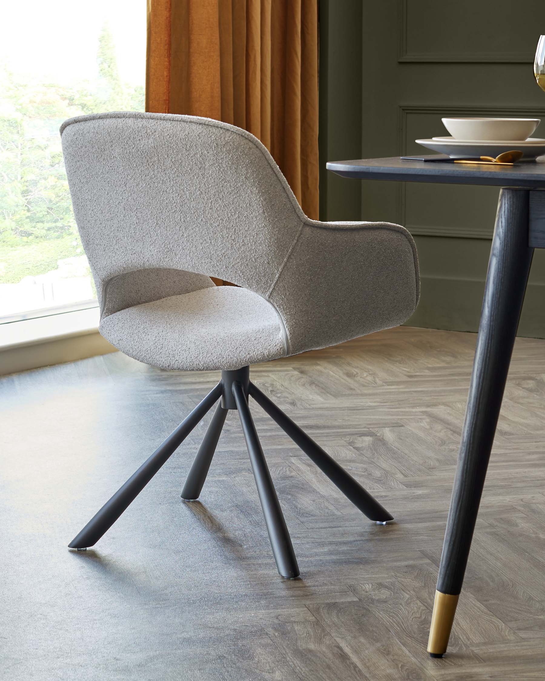 Modern upholstered chair with a curved backrest and cushioned seat in light grey fabric, featuring a swivel base with angled black metal legs tipped with brass accents, paired with a round-cornered dark blue table with slender black legs.