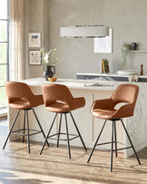 Three modern caramel-coloured upholstered bar stools with backrests and slender black metal legs arranged around a kitchen island.