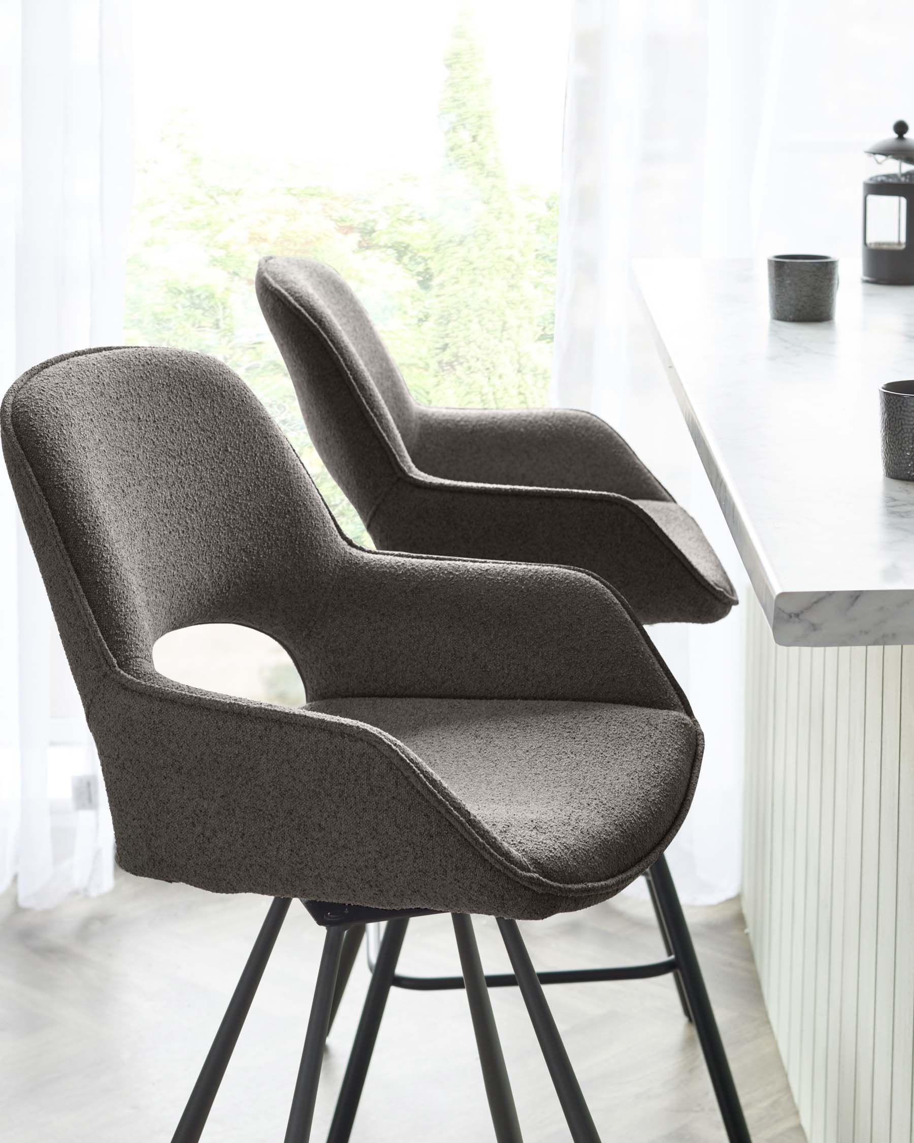 Two modern upholstered bar chairs with ergonomic armrests and textured dark grey fabric, positioned next to a white marble bar table accented with minimalist black vases. The chairs are supported by sleek black metal legs in a room with soft natural lighting.