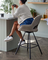 Modern textured fabric barstool with a curved high backrest, cushioned seat, and tapering matte black metal legs with a footrest.