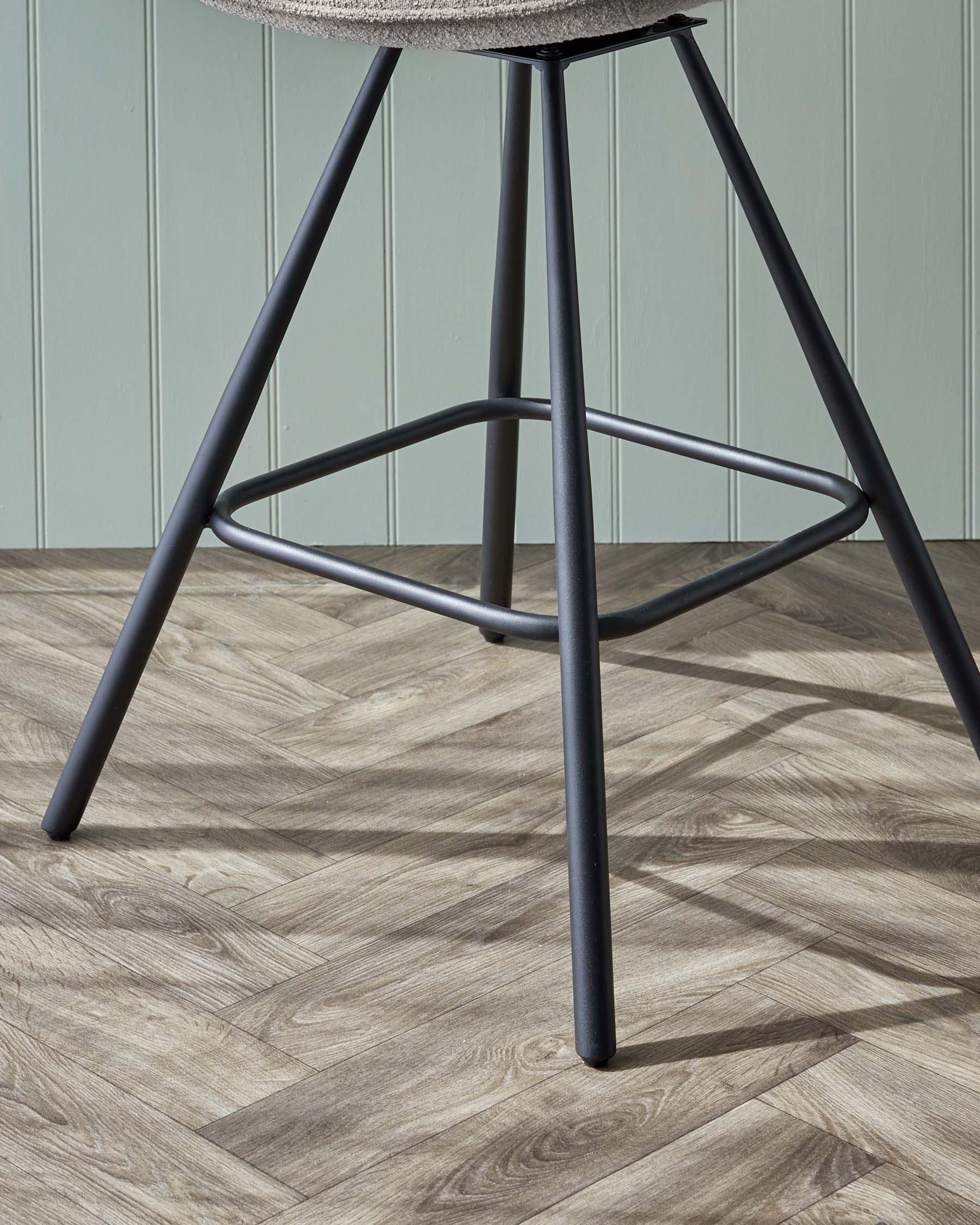 Modern stool with a grey fabric seat and sleek black metal legs in a triangular formation, showing a focus on minimalist design and stability, set against a wooden floor and pale green wall background.