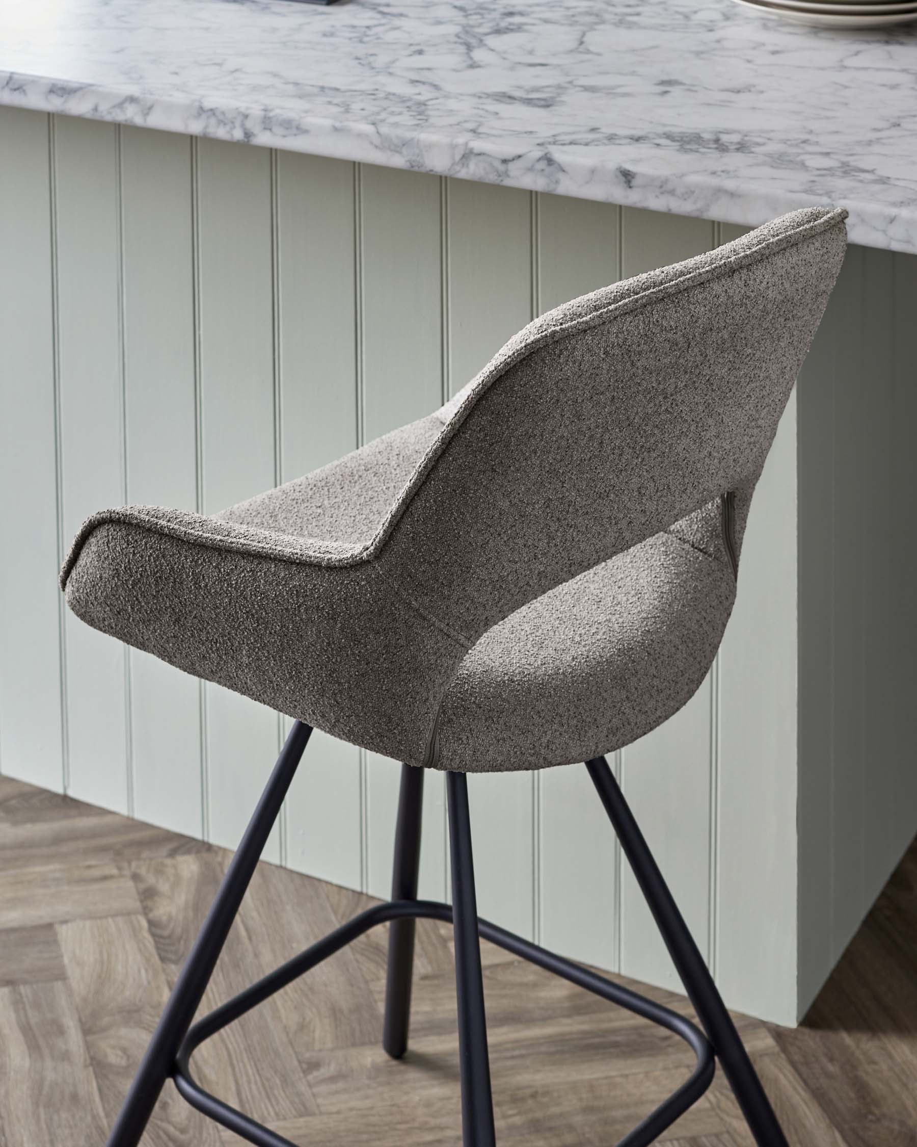 A modern upholstered bar stool with a textured grey fabric, featuring a curved backrest and a sleek black metal base with four legs.