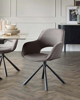 Modern upholstered dining chair with a seamless curved design and four splayed metal legs, positioned next to an elegant minimalist dining table with stacks of coordinating dinnerware on top. A sophisticated sideboard with textured front panels is visible in the background.