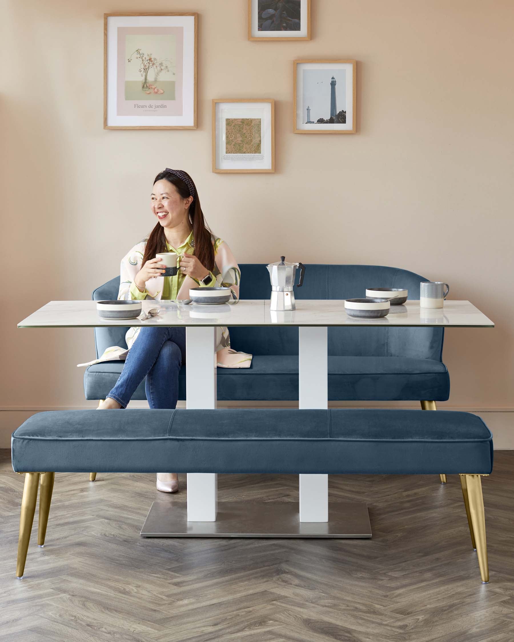 Modern dining set featuring a white rectangular table with sleek, chrome-finished central support and a base plate. Paired with a blue upholstered bench with plush seating and metallic gold-tipped legs.