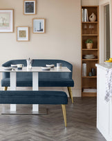 Modern dining room furniture consisting of a sleek oval table with a glossy white top and tapered metallic gold legs, paired with a matching curved bench-style seat upholstered in dark blue velvet with gold-toned legs. A wooden tall narrow bookshelf with graduated shelves stands in the background, displaying assorted decorative items and books.