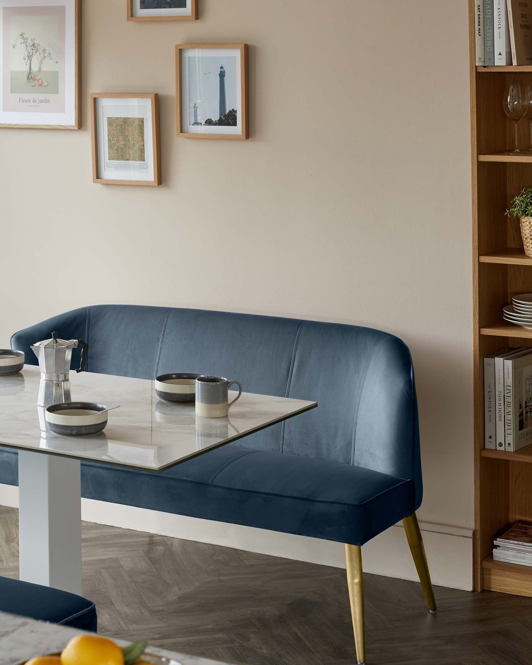 Elegant blue velvet half-circle dining bench with tapered gold legs, paired with a modern rectangular marble-top dining table with a unique blue and white central support. A tall wooden open shelving unit with assorted decorative items stands in the background.