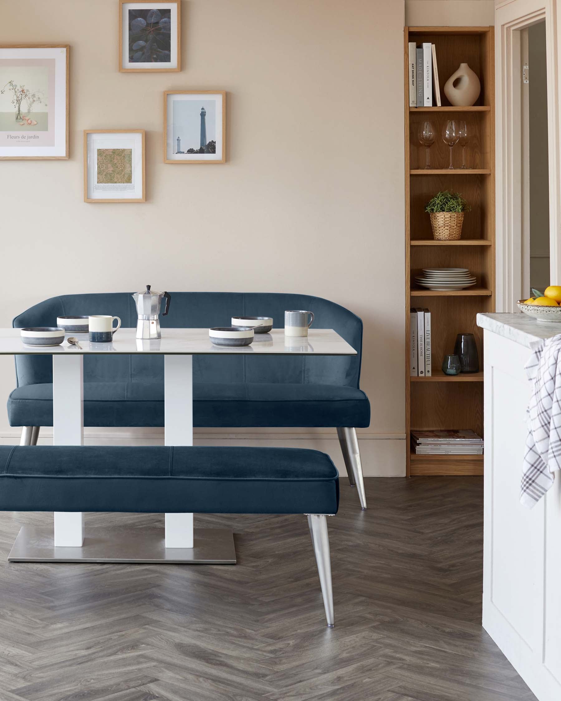 A modern dining set comprising a sleek rectangular table with a dark blue finish and contrasting white stripe accents sits atop a stainless steel base. Accompanying the table is a matching L-shaped corner bench and a separate bench, both upholstered in dark blue fabric with white support legs and a metallic base, offering a contemporary, minimalistic appeal. A tall, narrow wooden shelving unit, displaying a curated selection of books and decorative items, is situated against the wall.